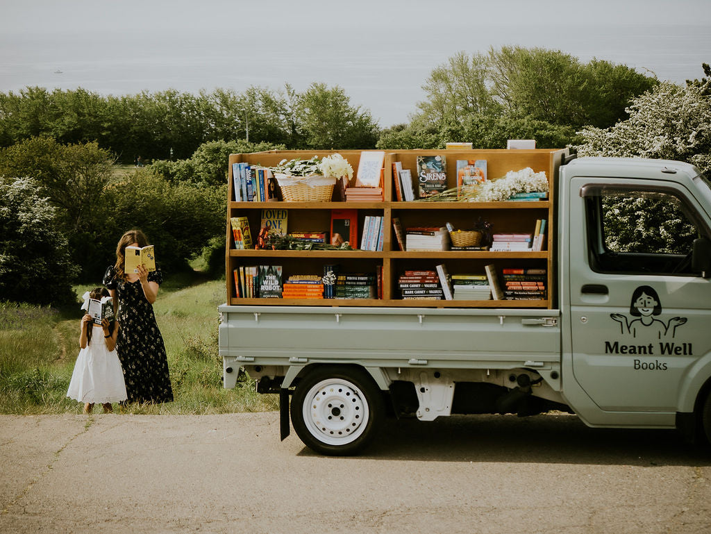 Two people reading beside the Meant Well Books truck, with bookshelves along its side.
